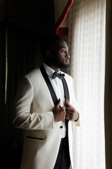groom in a white tuxedo with black collar posing for a photo at the window